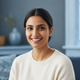 Headshot of a smiling South Asian woman with dark hair, looking at the camera with a warm expression. Blurred blue background